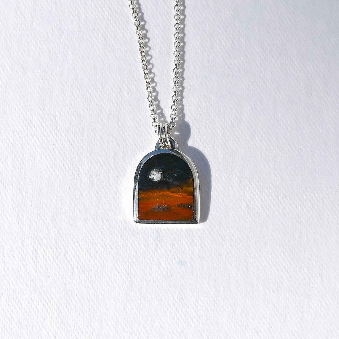 A silver chain necklace with an arched pendant featuring a sunset landscape design, displayed on a textured white background.