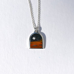 A silver chain necklace with an arched pendant featuring a sunset landscape design, displayed on a textured white background.