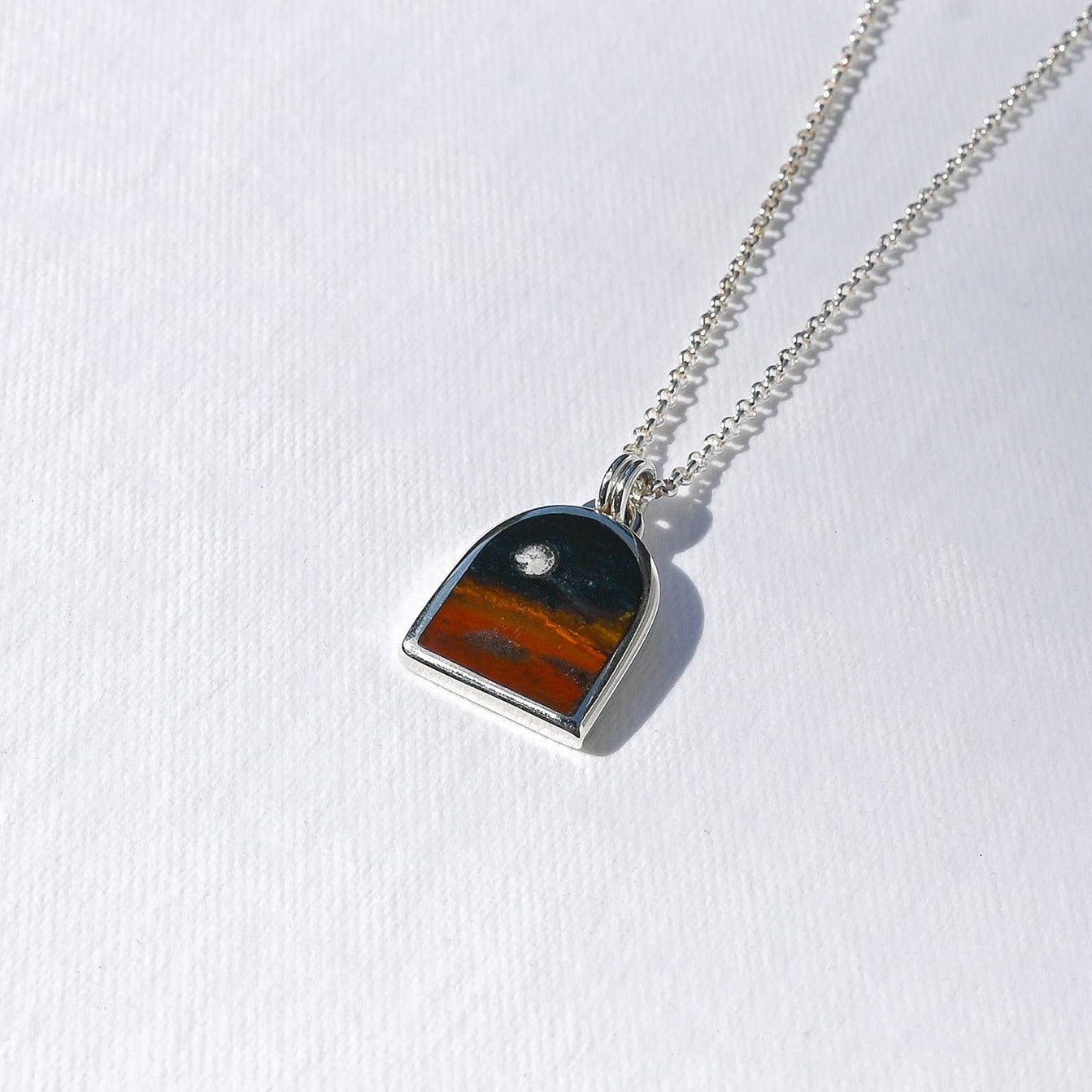 A silver chain necklace with a small arch-shaped pendant featuring a sunset landscape design, displayed on a textured white background.