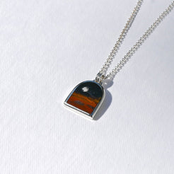 A silver chain necklace with a small arch-shaped pendant featuring a sunset landscape design, displayed on a textured white background.