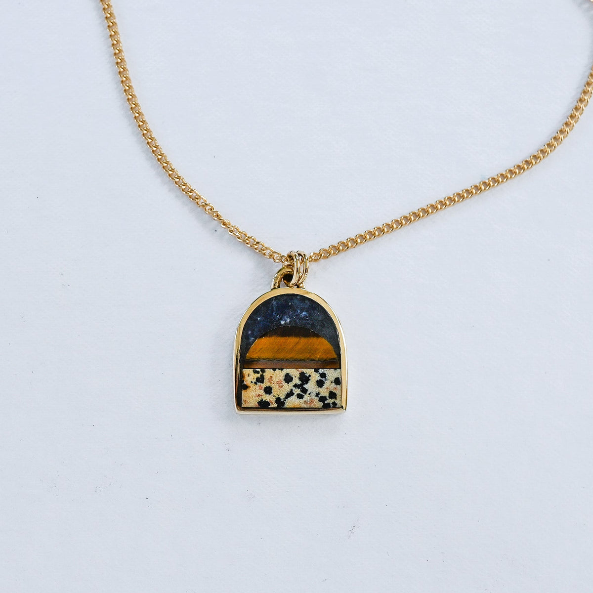 A gold chain necklace with an arched pendant featuring a layered inlay design: a dark navy lapis blue top section, a warm tiger eye middle band, and a dalmatian stone lower section with black spots, all set in a gold frame on a white background.