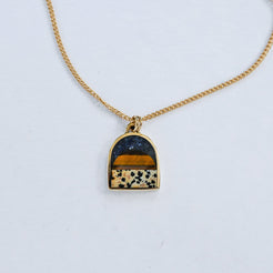 A gold chain necklace with an arched pendant featuring a layered inlay design: a dark navy lapis blue top section, a warm tiger eye middle band, and a dalmatian stone lower section with black spots, all set in a gold frame on a white background.