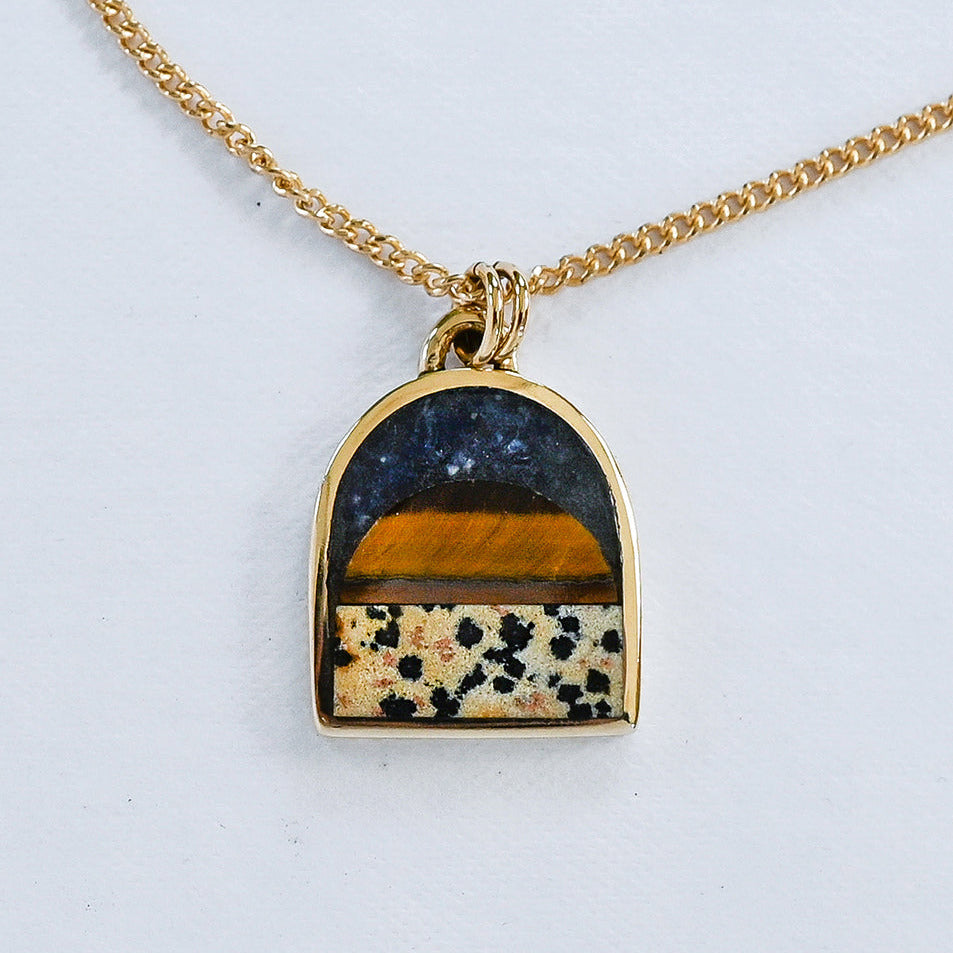 A gold chain necklace with an arched pendant featuring a layered inlay design: a dark navy lapis blue top section, a warm tiger eye middle band, and a dalmatian stone lower section with black spots, all set in a gold frame on a white background.
