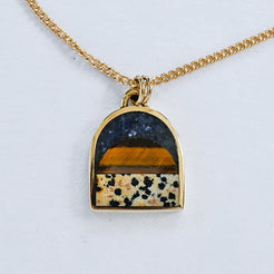 A gold chain necklace with an arched pendant featuring a layered inlay design: a dark navy lapis blue top section, a warm tiger eye middle band, and a dalmatian stone lower section with black spots, all set in a gold frame on a white background.