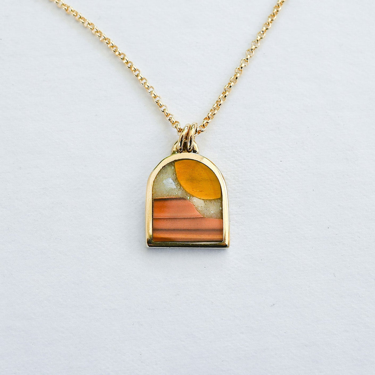 Gold necklace with a small arch-shaped pendant featuring a stone inlay design of tiger eye sun, orange and brown wave dolomite with a cream colored mother of pearl sky.