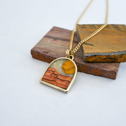 Gold pendant necklace with a landscape inlay featuring a striped tiger eye sun, a dune-like orange wave dolomite landscape and a pale mother of pearl sky, displayed on slabs of natural stone