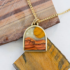 Gold pendant necklace with a landscape inlay featuring a striped tiger eye sun, a dune-like orange wave dolomite landscape and a pale mother of pearl sky, displayed on slabs of natural stone