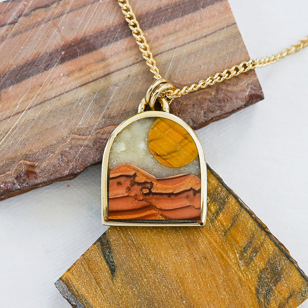 Gold pendant necklace with a landscape inlay featuring a striped tiger eye sun, a dune-like orange wave dolomite landscape and a pale mother of pearl sky, displayed on slabs of natural stone