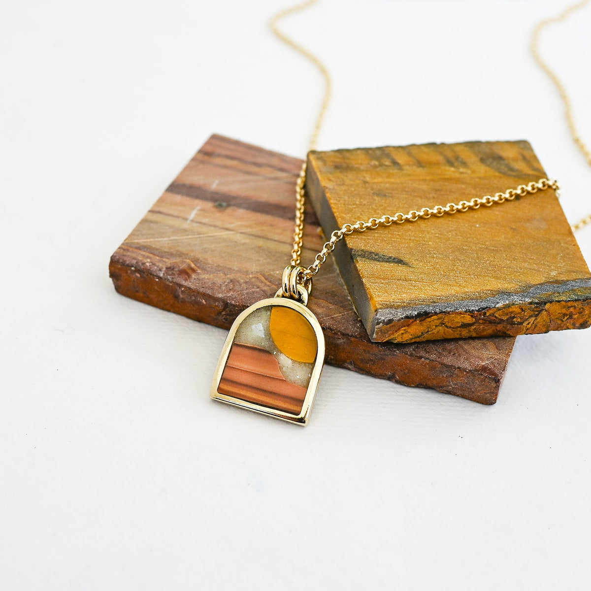 Gold pendant necklace with a landscape inlay featuring a striped tiger eye sun, a dune-like orange wave dolomite landscape and a pale mother of pearl sky, displayed on slabs of natural stone