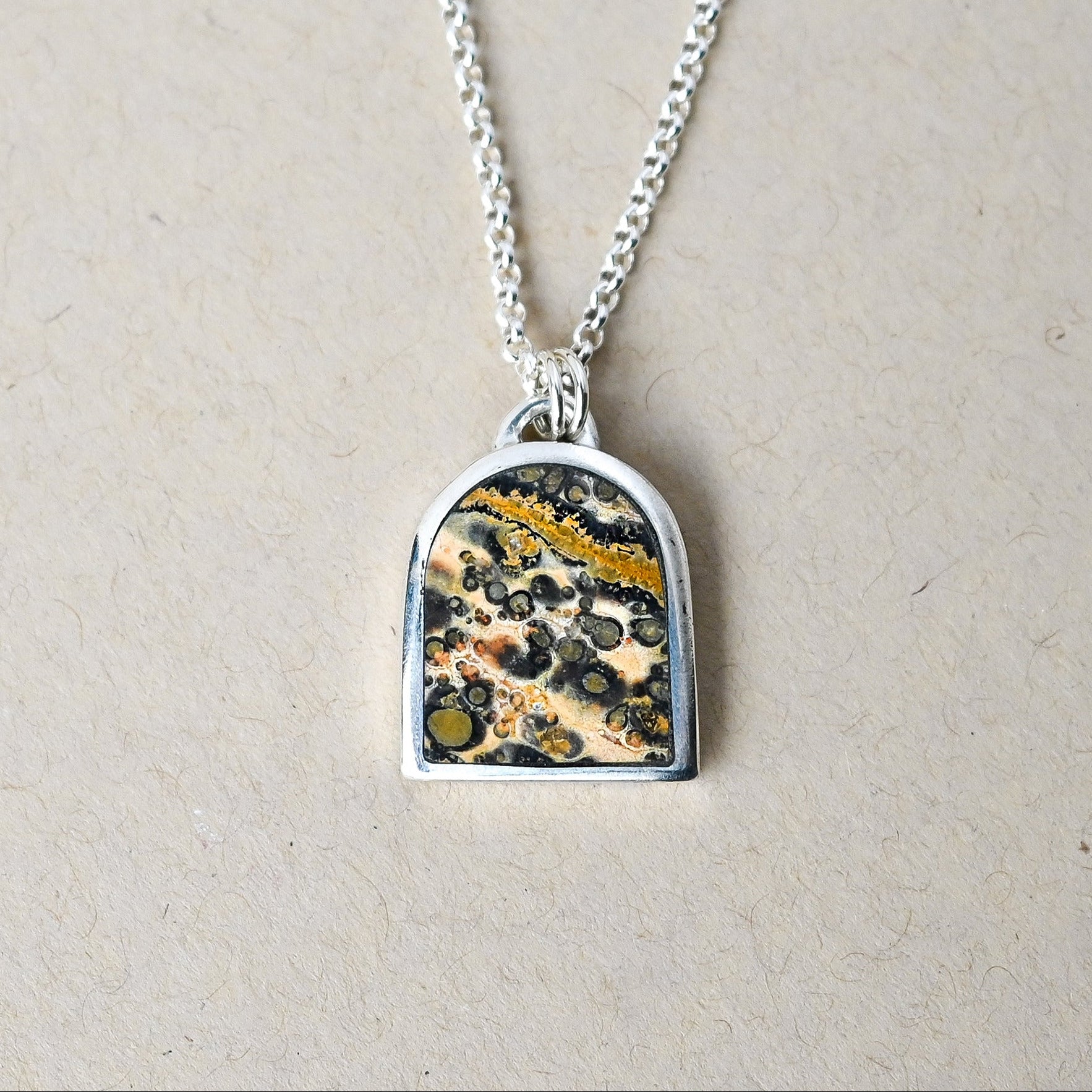 sterling silver necklace with an arch shaped pendant featuring leopard skin jasper stone on a beige background
