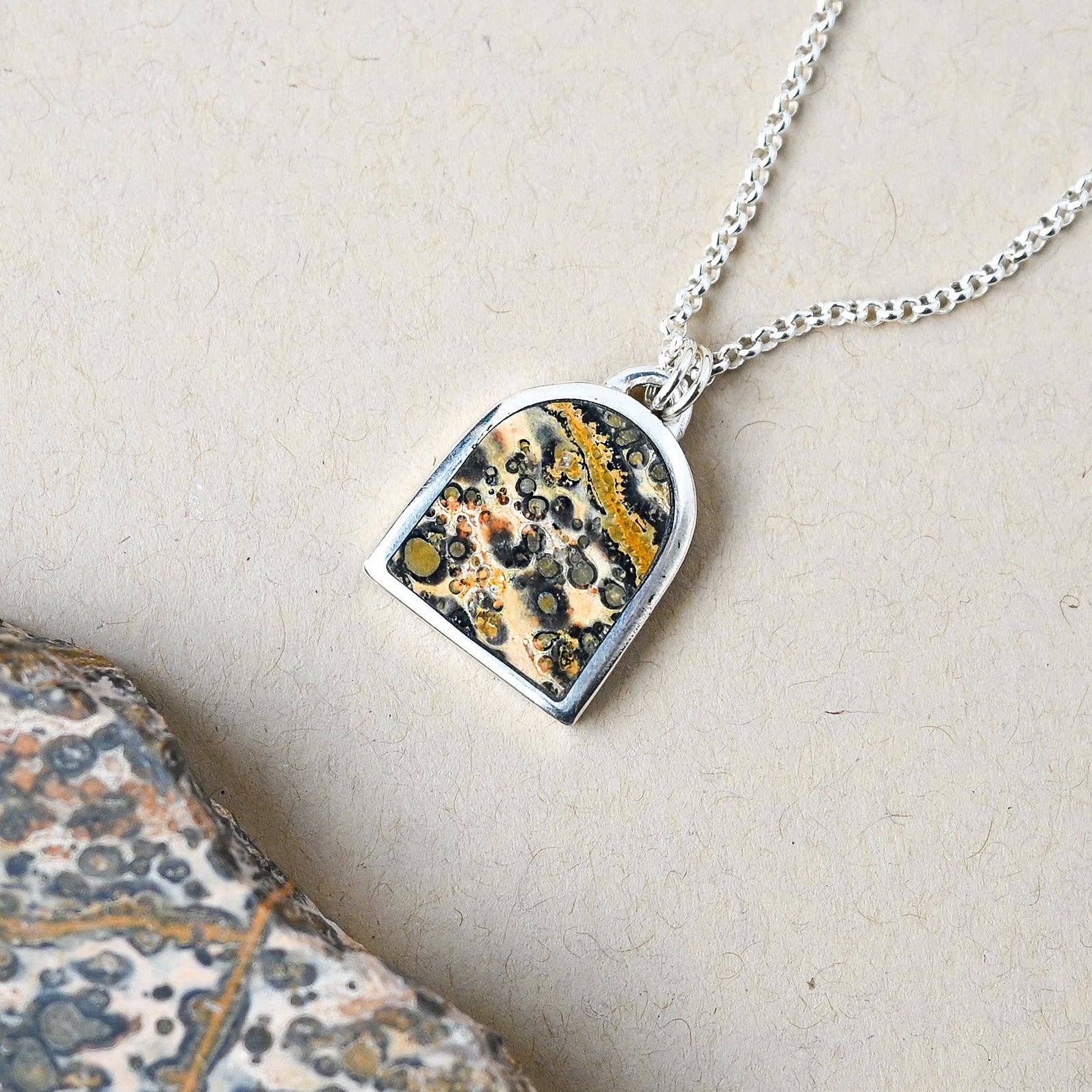 sterling silver necklace with an arch shaped pendant featuring leopard skin jasper stone on a beige background