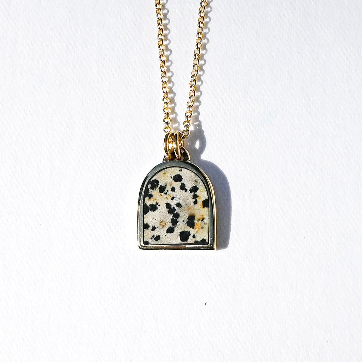A gold chain necklace with a small arch-shaped pendant featuring a white stone inlay speckled with black and gold flecks, set against a plain white background.