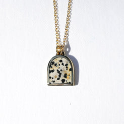 A gold chain necklace with a small arch-shaped pendant featuring a white stone inlay speckled with black and gold flecks, set against a plain white background.