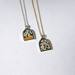 Two pendant necklaces on white textured paper. One necklace has a silver chain and a pendant with a black and white speckled pattern and a yellow stripe.