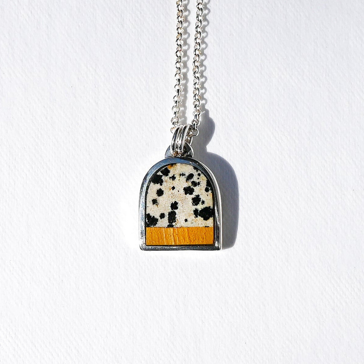 A silver chain necklace with an arched pendant featuring a black-and-white cow print top and a textured orange bottom, displayed on a white background.