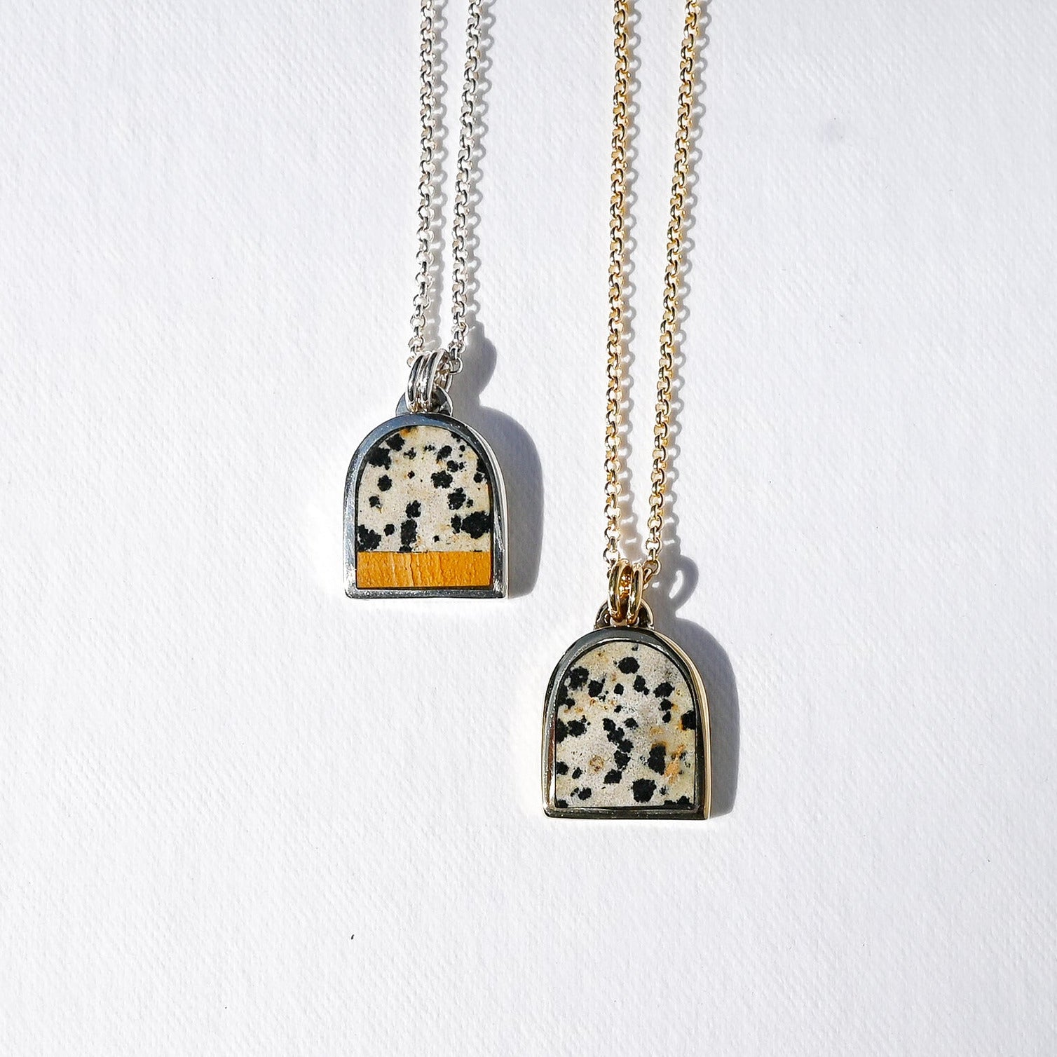 Two arched pendant necklaces hanging against a white textured background. One necklace has a silver chain and a pendant with a black-and-white speckled pattern and a yellow-orange accent at the bottom.