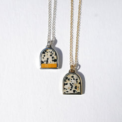 Two arched pendant necklaces hanging against a white textured background. One necklace has a silver chain and a pendant with a black-and-white speckled pattern and a yellow-orange accent at the bottom.