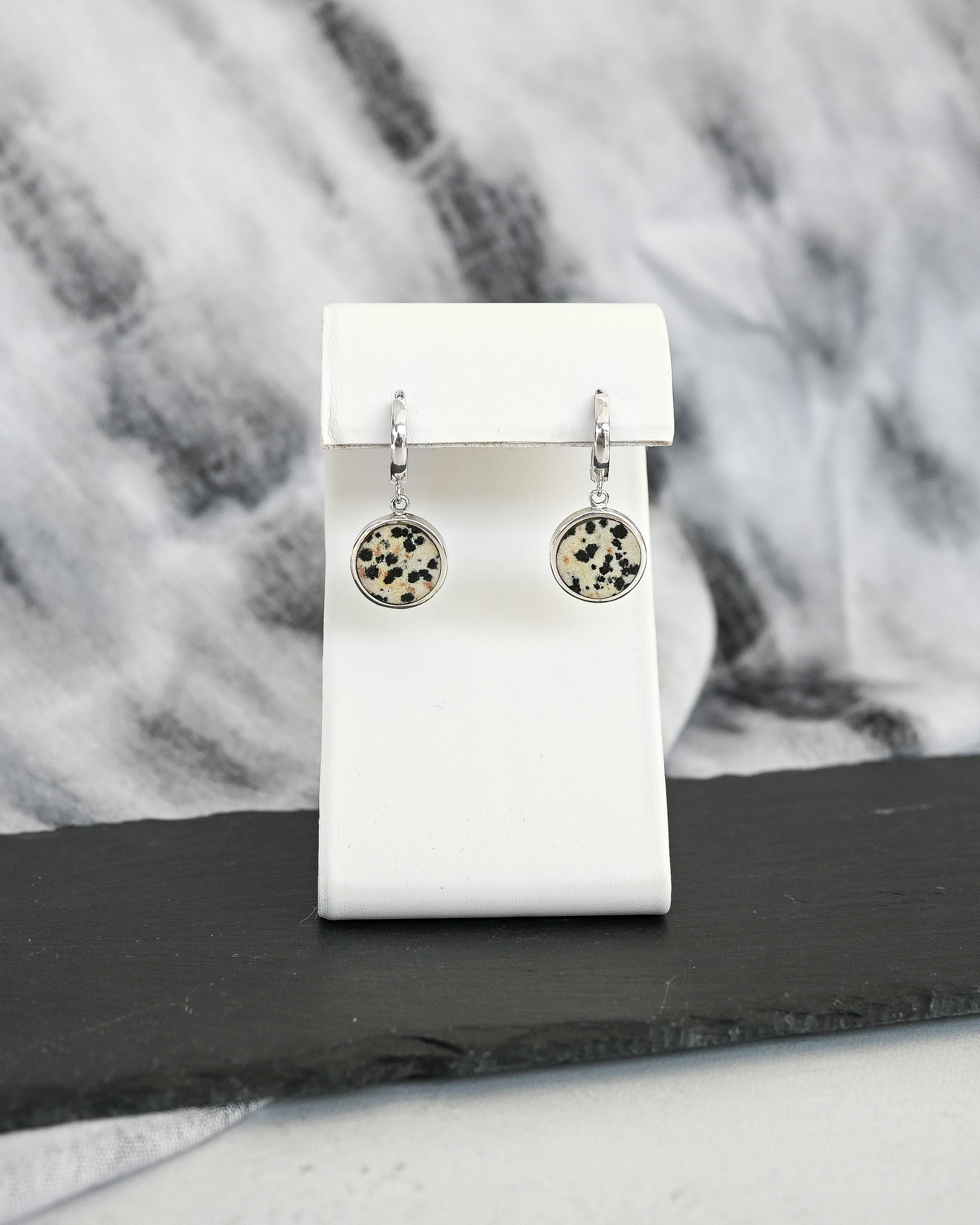 Pair of silver earrings with circular designs featuring dalmatian stone on a white card against a fabric background