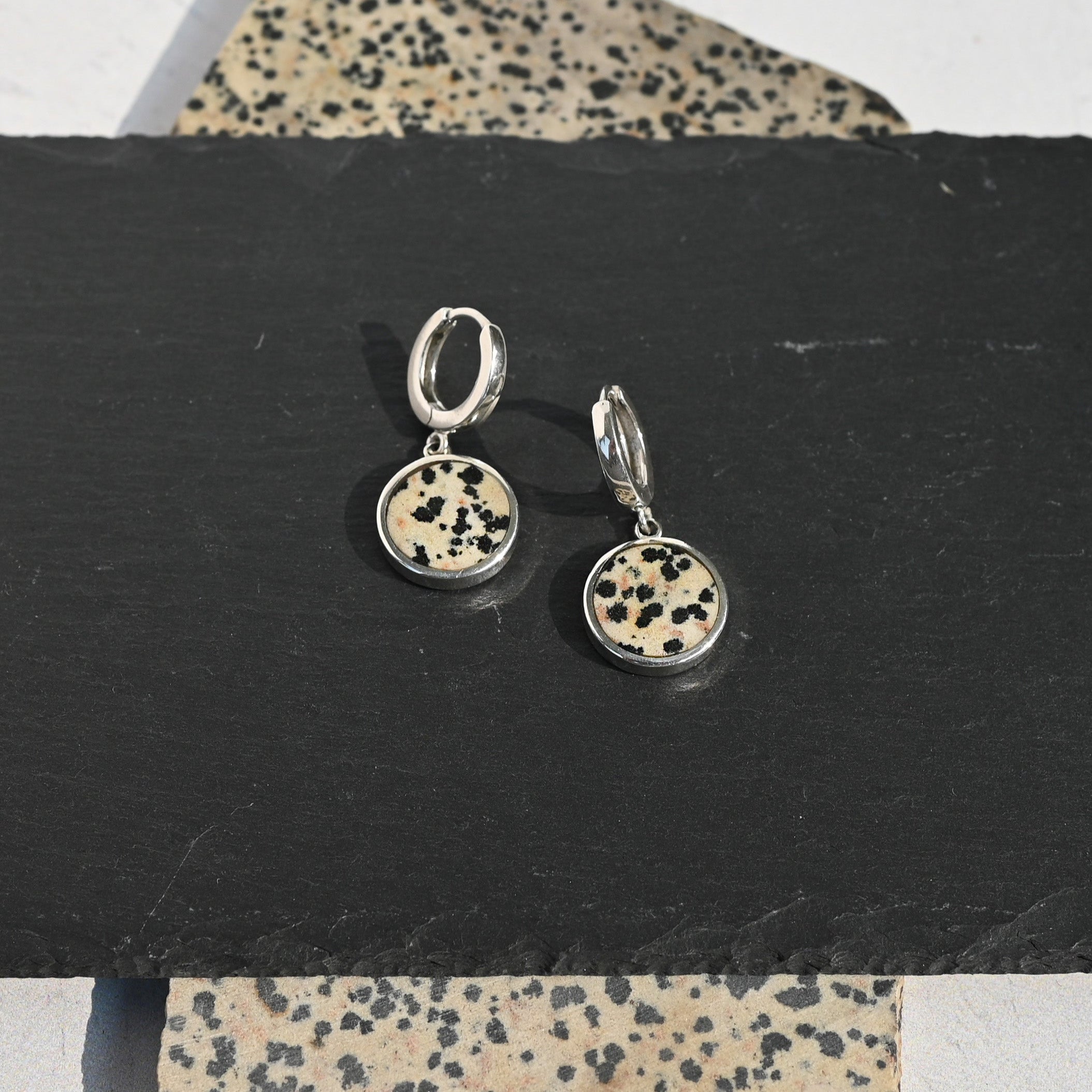 Pair of sterling silver huggie earrings with dalmatian stone charms on a black stand with a patterned surface underneath.
