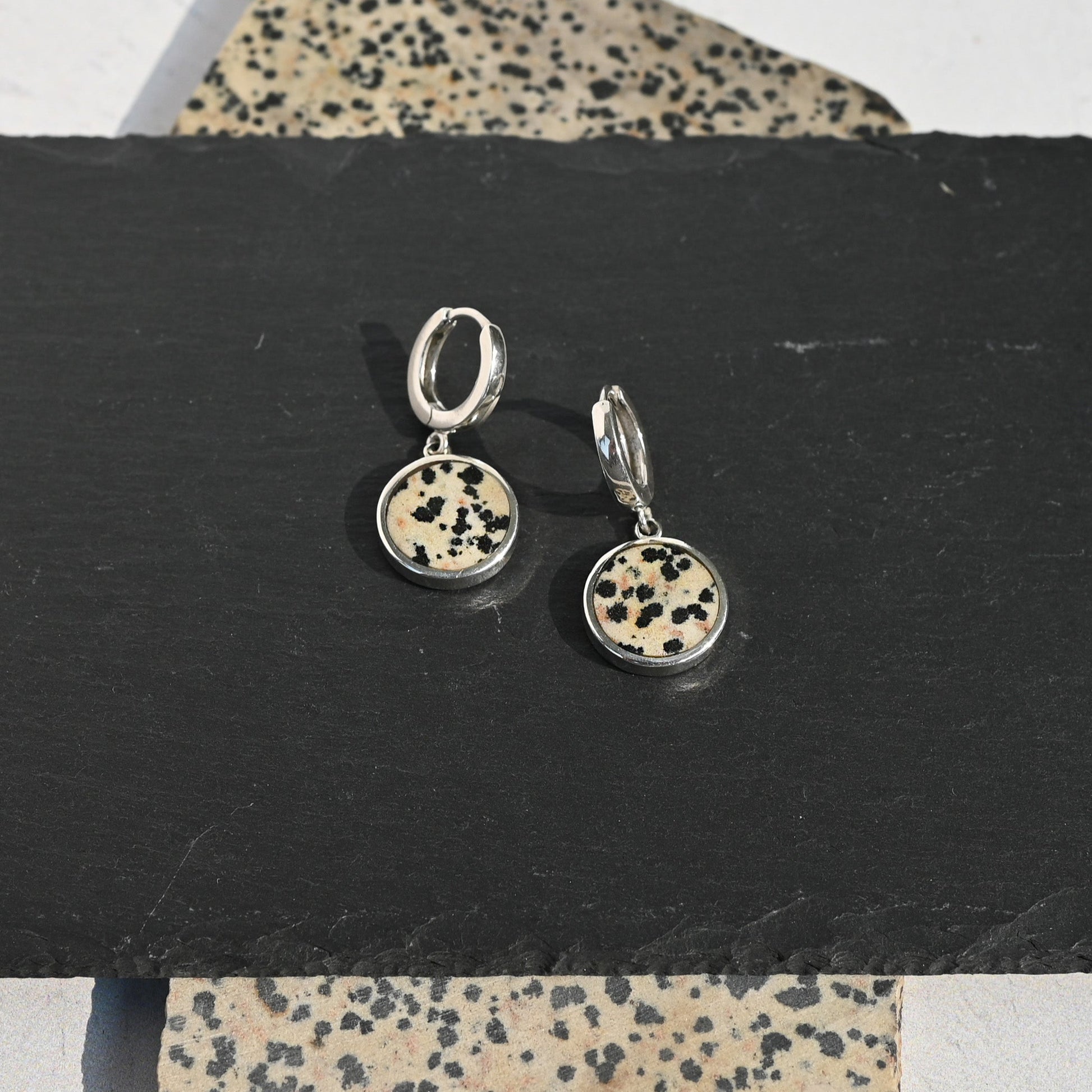 Pair of sterling silver huggie earrings with dalmatian stone charms on a black stand with a patterned surface underneath.