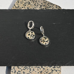 Pair of sterling silver huggie earrings with dalmatian stone charms on a black stand with a patterned surface underneath.