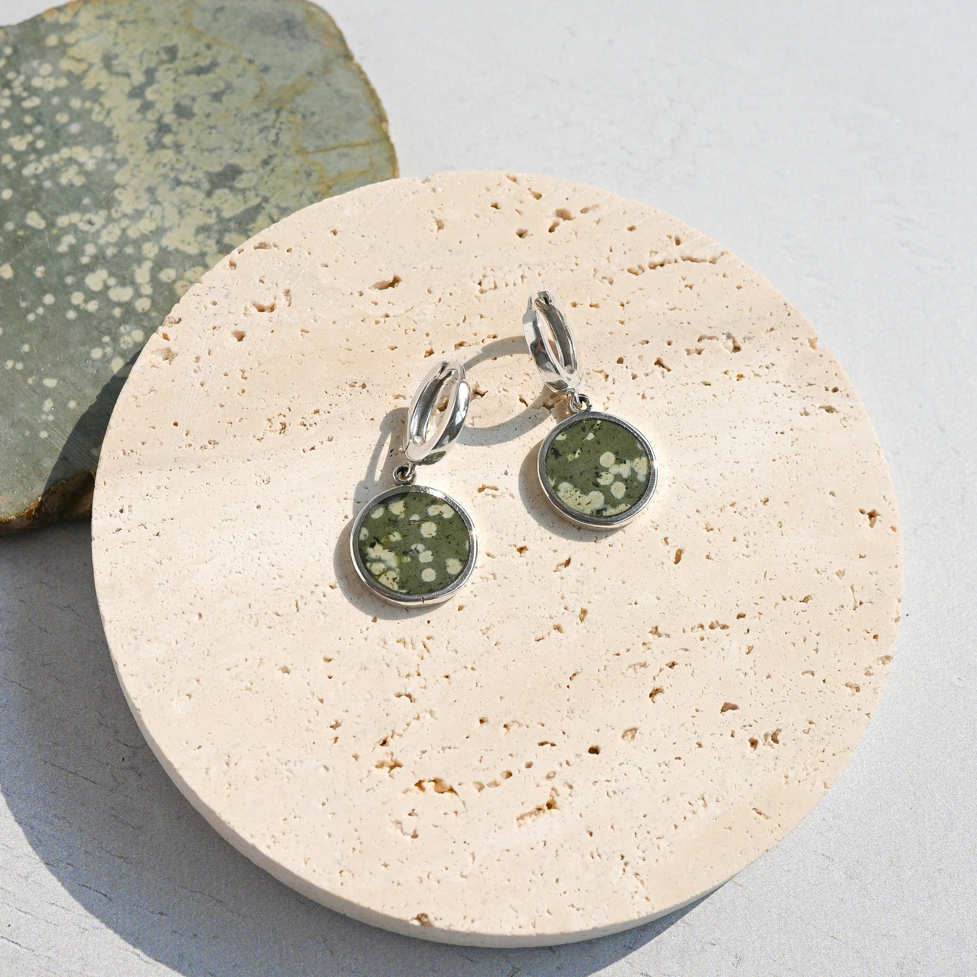 Pair of sterling silver huggie earrings with green variolite stone charms on a stone surface
