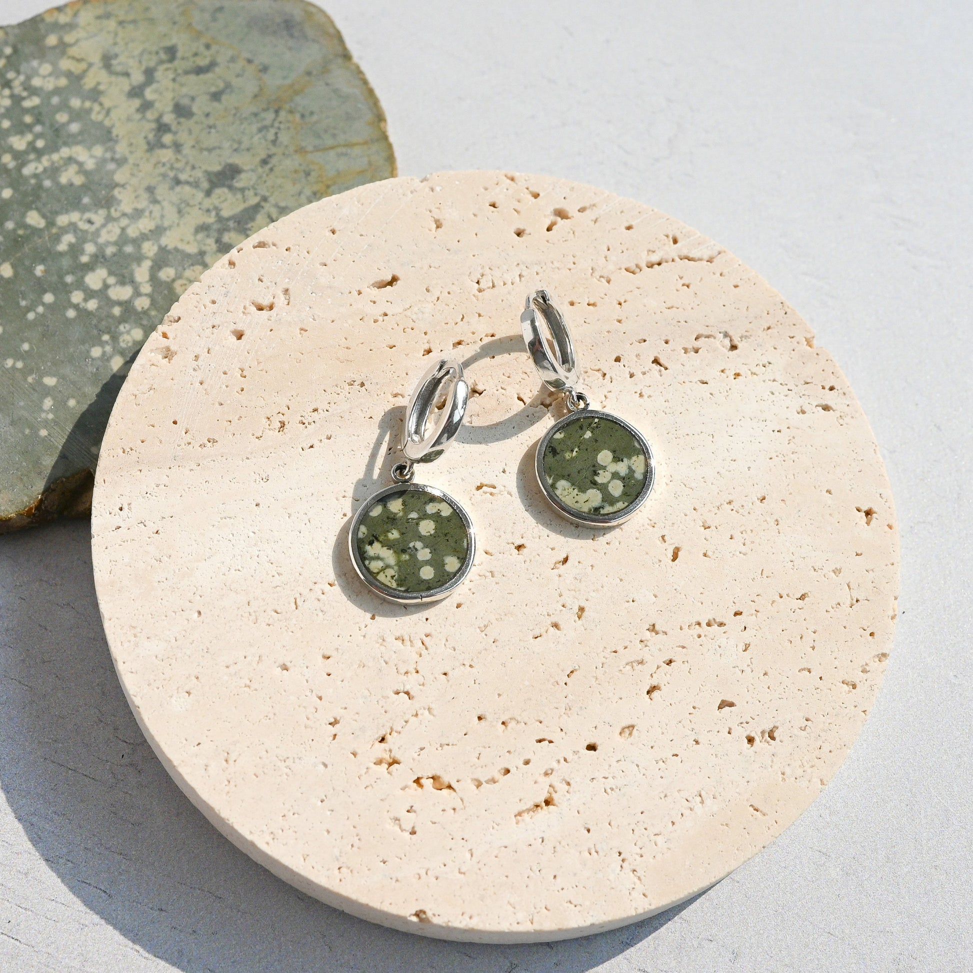 Pair of sterling silver huggie earrings with green variolite stone charms on a stone surface