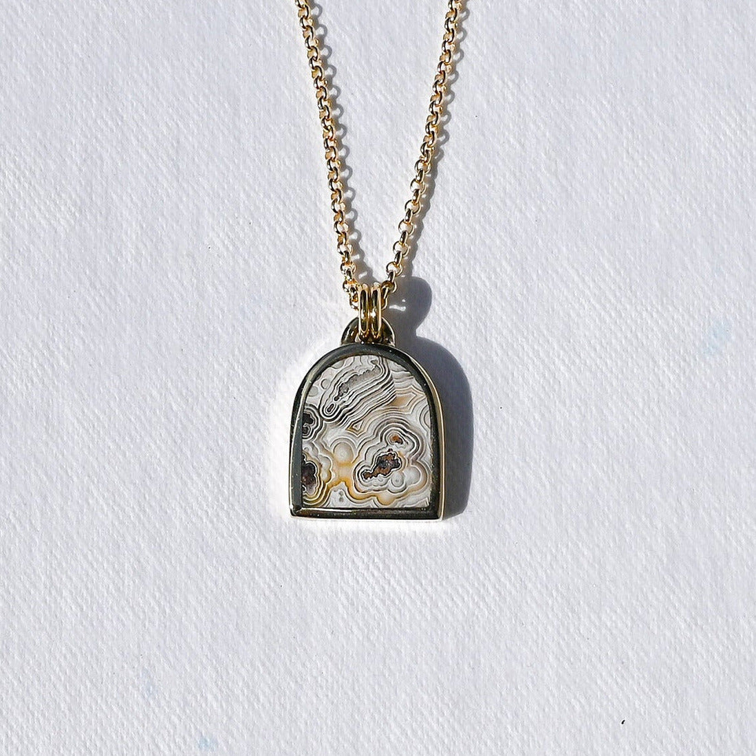 A gold chain necklace with a small arched pendant featuring a white and gray patterned insert, photographed against a textured white background.