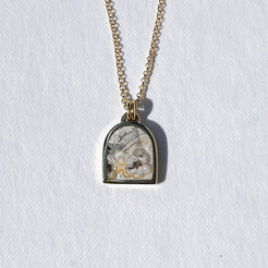 A gold chain necklace with a small arched pendant featuring a white and gray patterned insert, photographed against a textured white background.