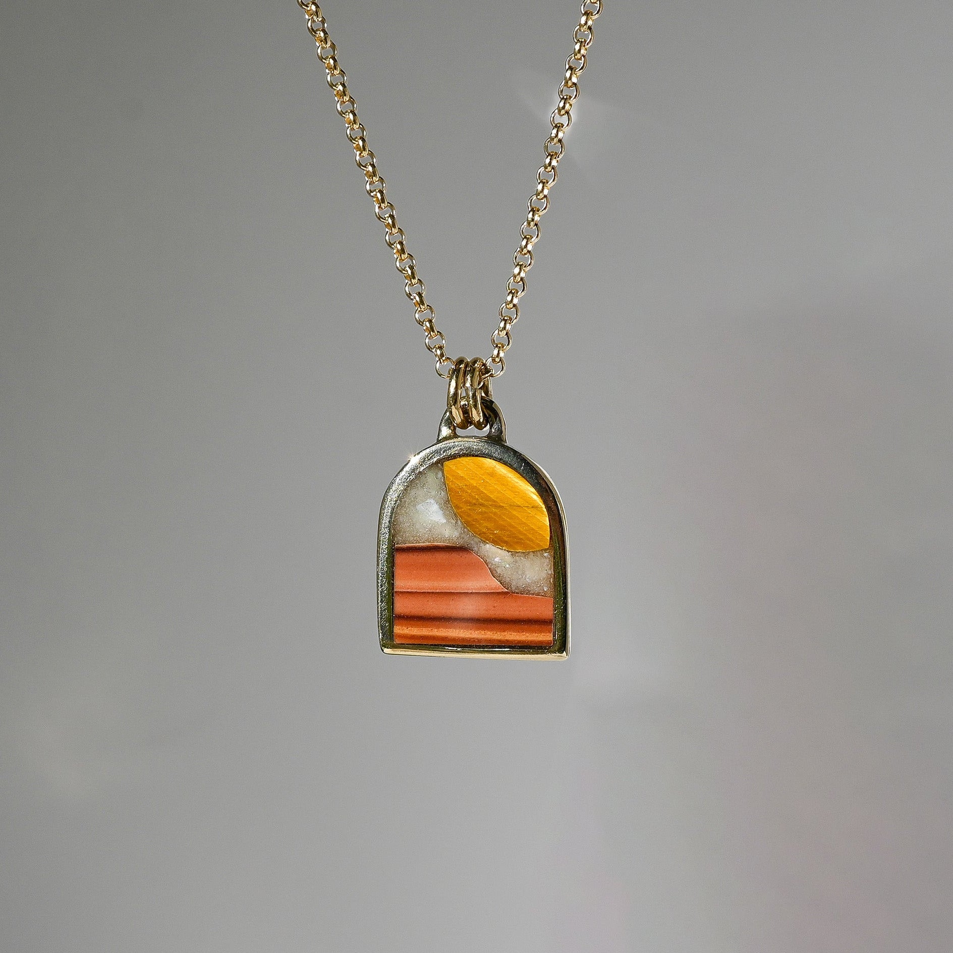 A gold chain necklace with a small arched pendant featuring a colorful inlay of orange, yellow, and white, set against a plain gray background.