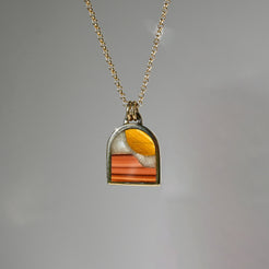 A gold chain necklace with a small arched pendant featuring a colorful inlay of orange, yellow, and white, set against a plain gray background.