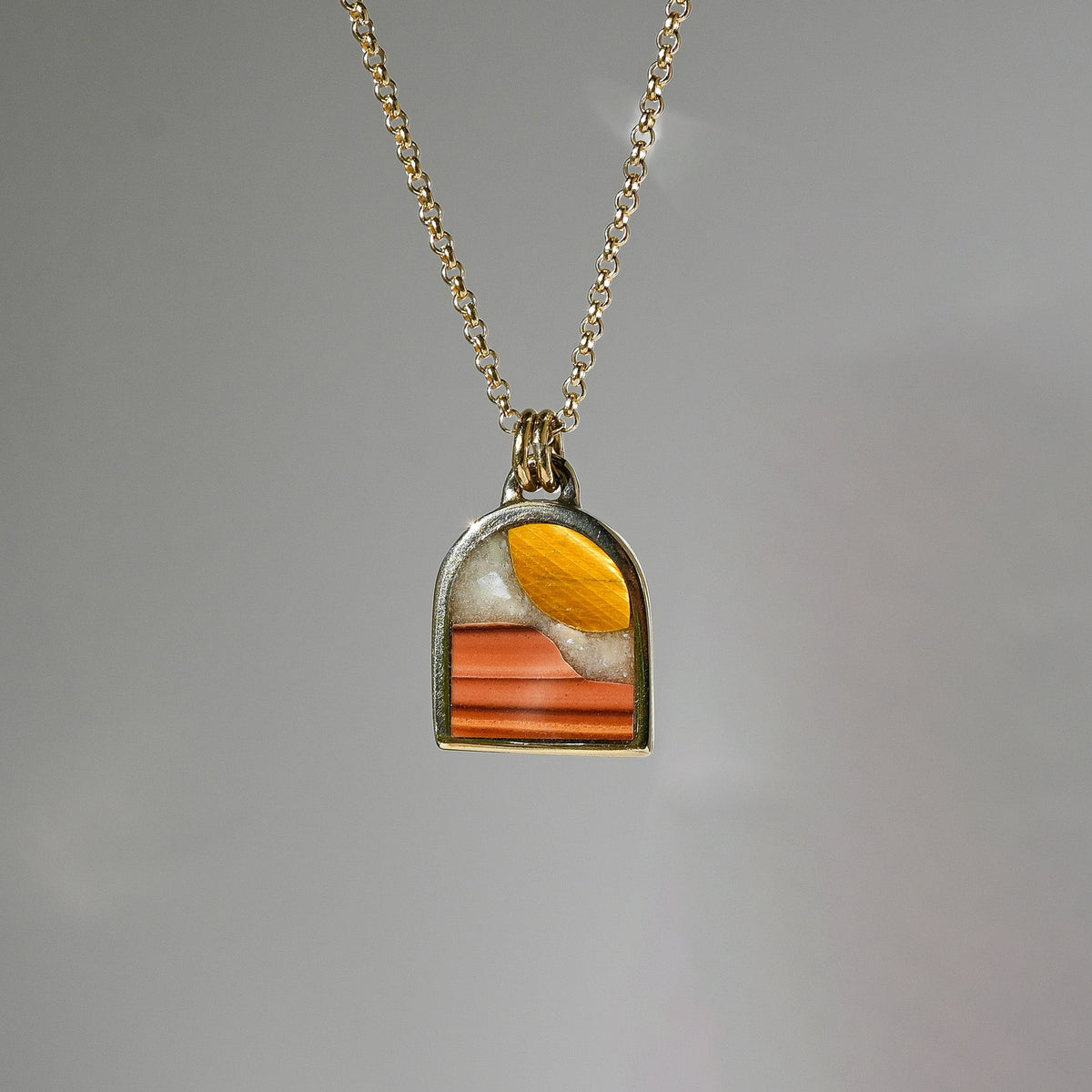 A gold chain necklace with a small arched pendant featuring a colorful inlay of orange, yellow, and white, set against a plain gray background.