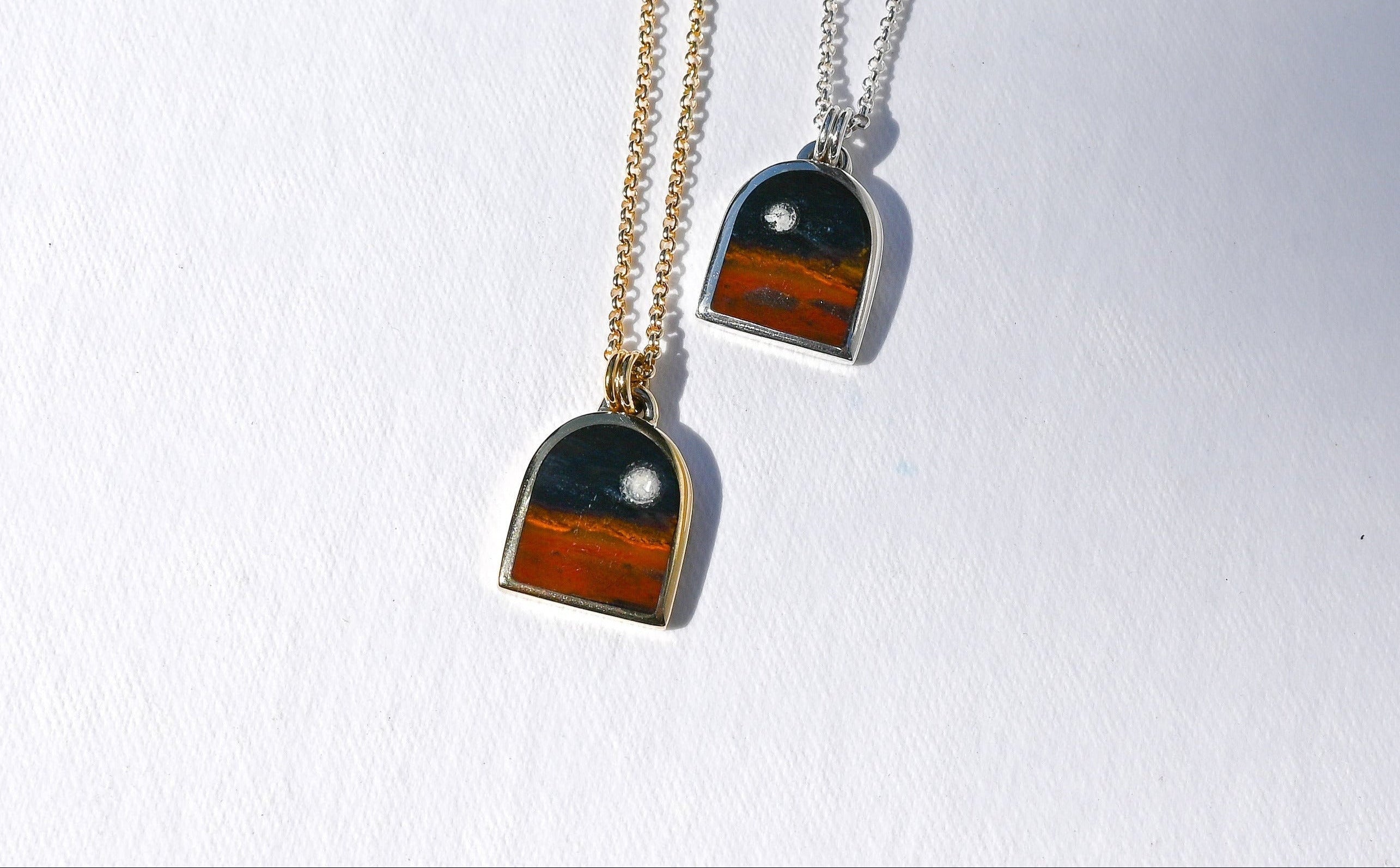 Two dome-shaped pendant necklaces on chains, one gold-toned and one silver-toned, featuring a layered design with dark gray, black, and orange-brown sections, displayed on a white textured surface.