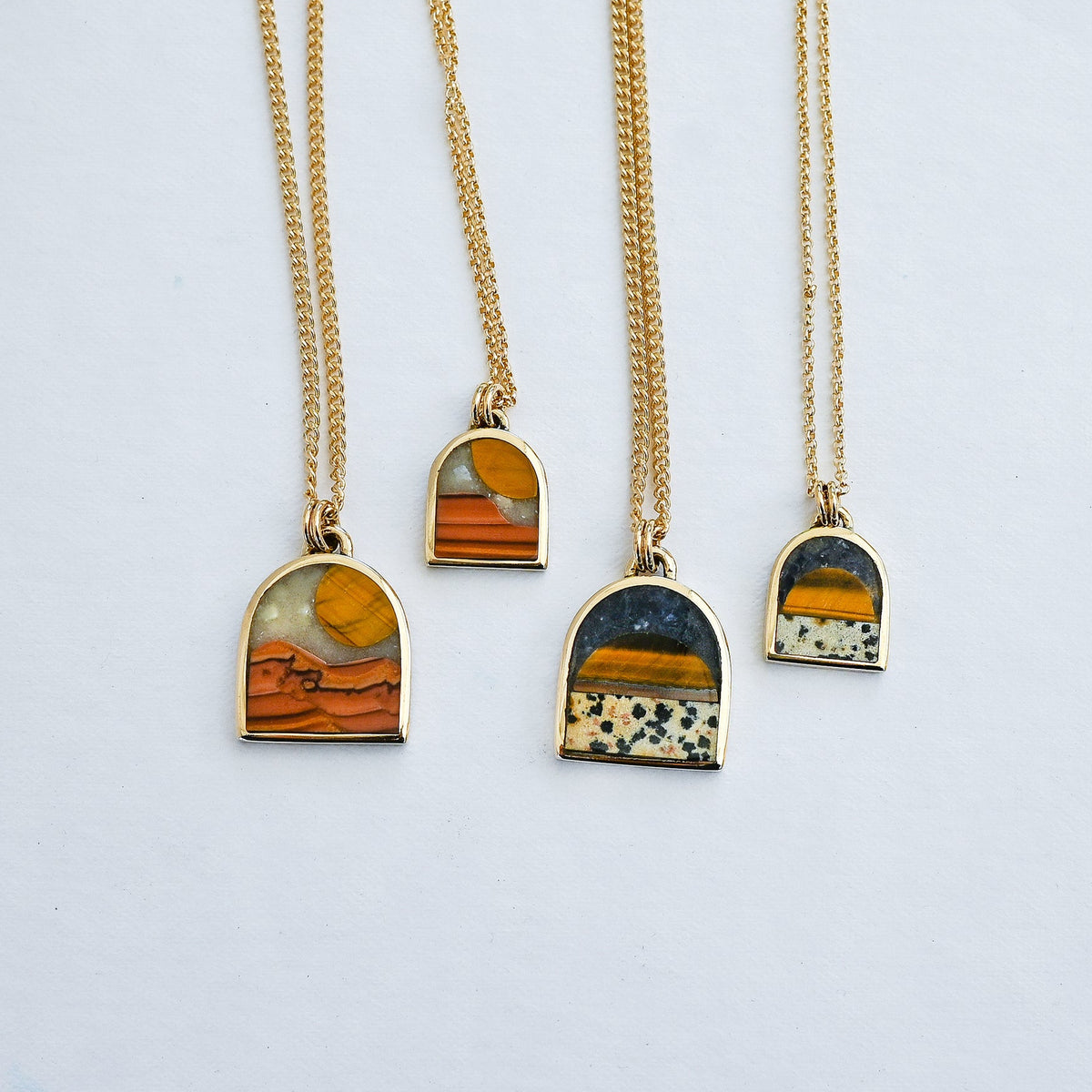 Four gold chain necklaces with arched pendant frames featuring natural stone inlays in orange, yellow, black, and white, arranged on a light gray background.