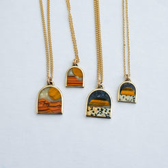 Four gold chain necklaces with arched pendant frames featuring natural stone inlays in orange, yellow, black, and white, arranged on a light gray background.