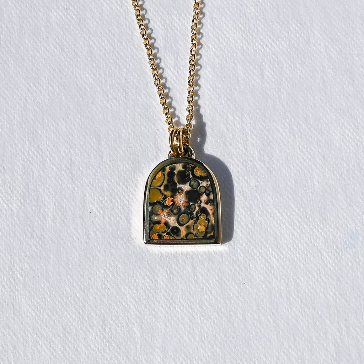 A gold chain necklace with an arched pendant featuring a colorful, textured mosaic design in yellow, green, and black, displayed on a textured white background.