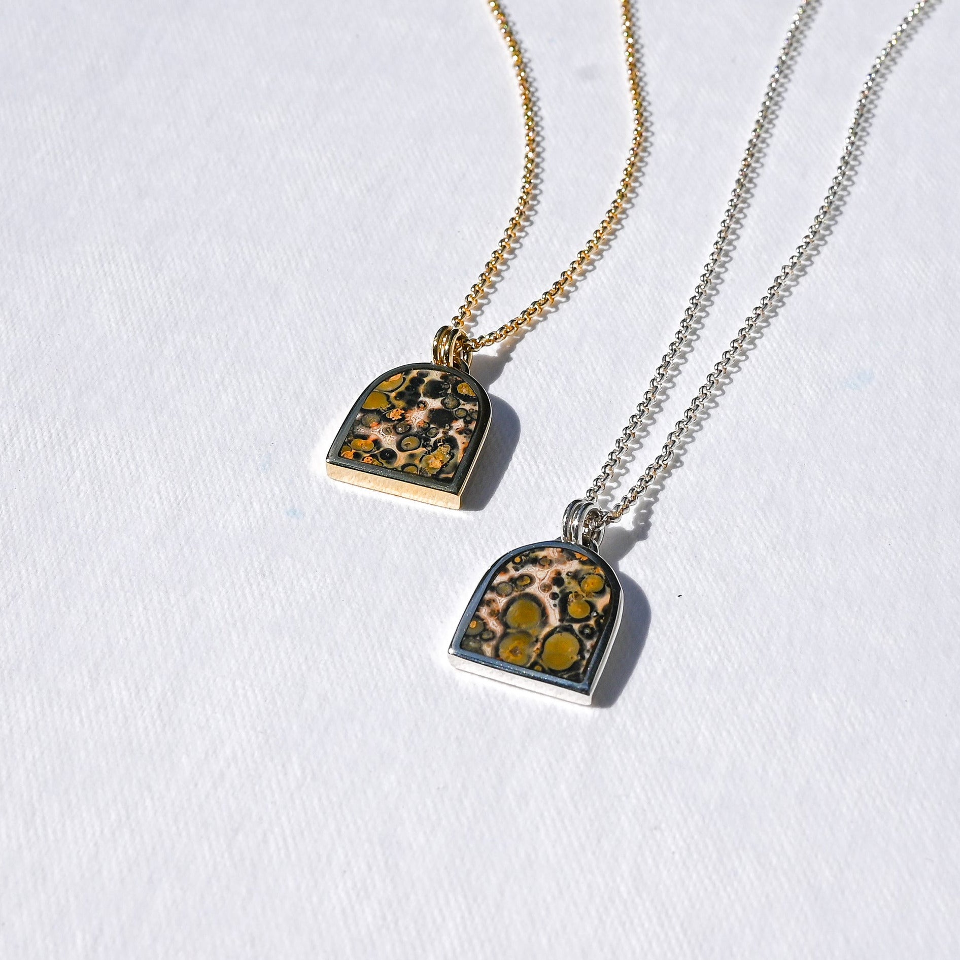 Two small pendant necklaces on white textured fabric, one with a gold chain and one with a silver chain. The arched square pendants feature a natural bird's-eye pattern with green, yellow, and black spots on a beige background.