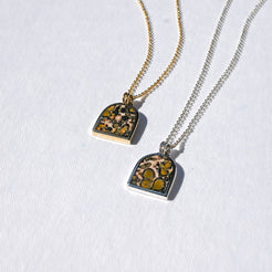 Two small pendant necklaces on white textured fabric, one with a gold chain and one with a silver chain. The arched square pendants feature a natural bird's-eye pattern with green, yellow, and black spots on a beige background.
