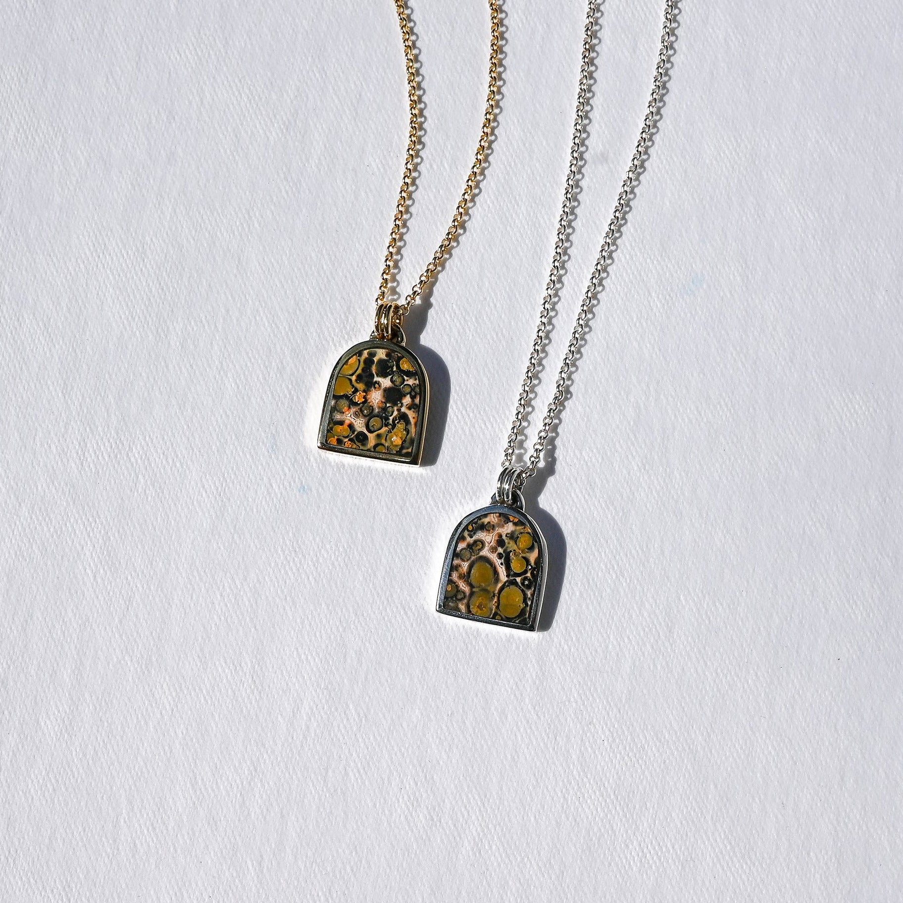 Two tombstone-shaped pendant necklaces on chains, one gold-toned and one silver-toned, featuring a yellow and black marbled pattern, displayed on a textured white background.
