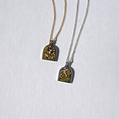 Two tombstone-shaped pendant necklaces on chains, one gold-toned and one silver-toned, featuring a yellow and black marbled pattern, displayed on a textured white background.
