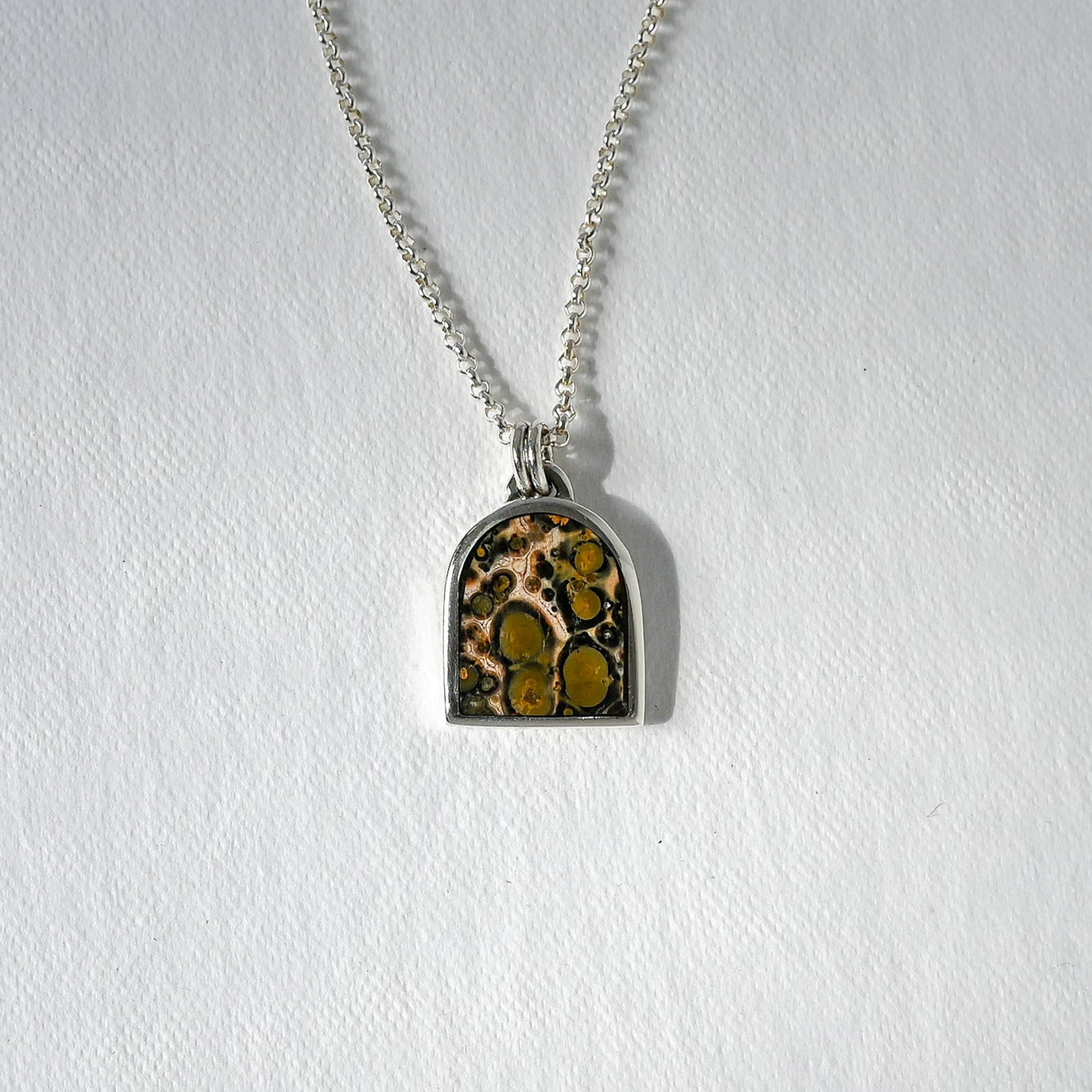 A silver chain necklace with an arched pendant featuring a brown and gold mottled pattern, displayed on a textured white background.