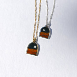 Two arched pendant necklaces on white textured paper. One necklace has a gold chain and a pendant with a black top section and a rust-orange bottom section.