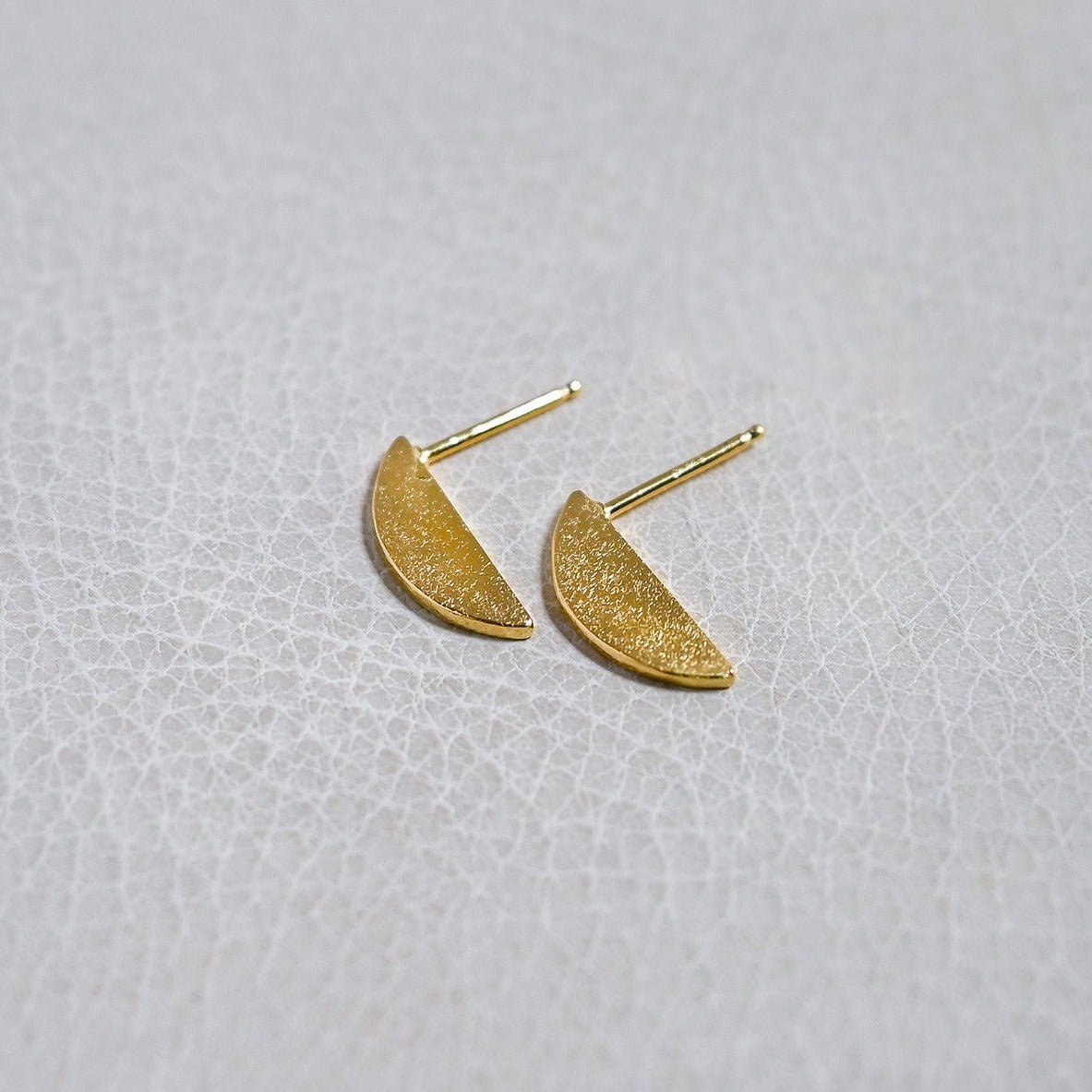 A pair of small gold crescent-shaped stud earrings on a textured light gray surface.