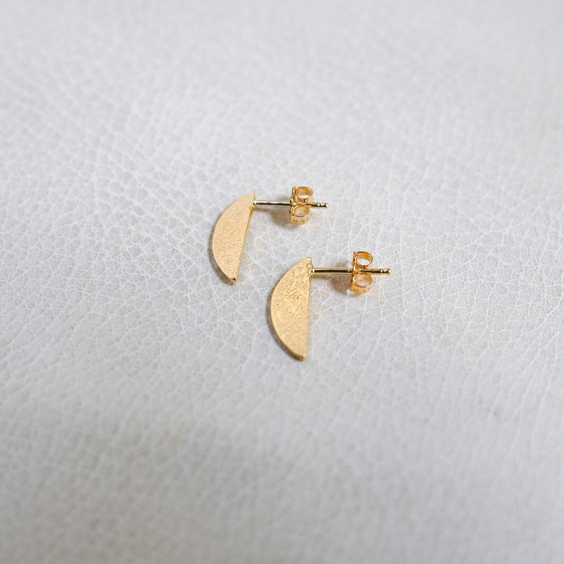 A pair of small gold crescent moon stud earrings on a textured white surface.