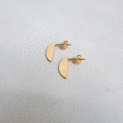 A pair of small gold crescent moon stud earrings on a textured white surface.