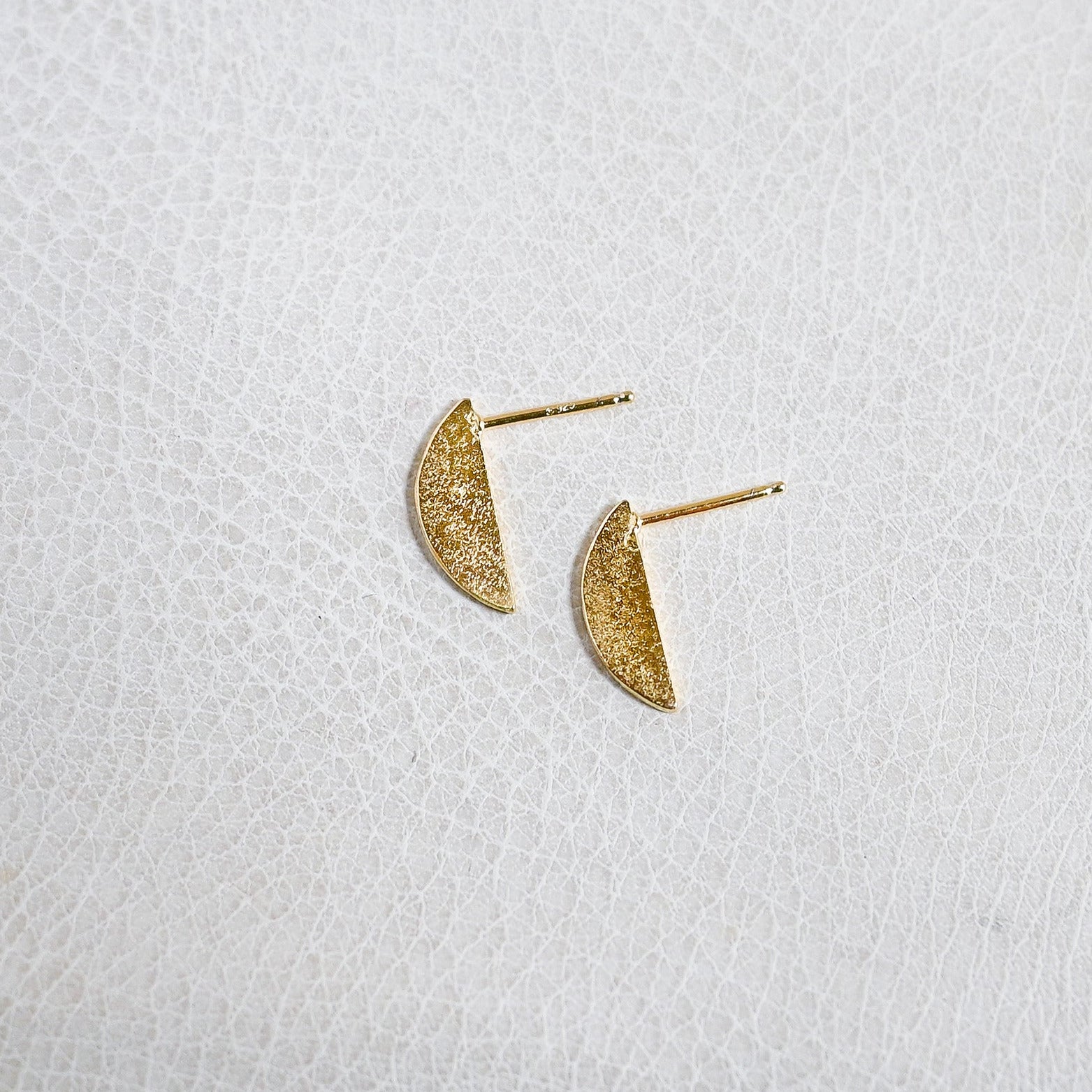 A pair of small gold crescent moon stud earrings with a textured, glittery finish, displayed on a white textured surface.