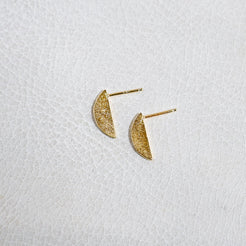 A pair of small gold crescent moon stud earrings with a textured, glittery finish, displayed on a white textured surface.