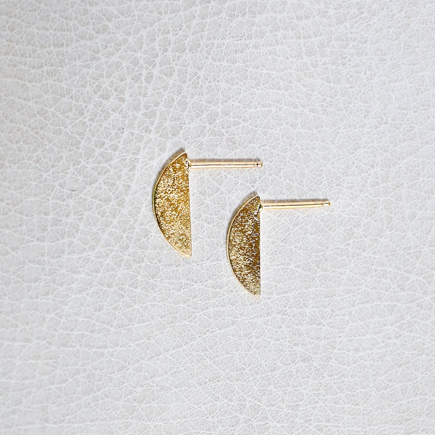 A pair of gold crescent-shaped stud earrings with a textured glitter finish, displayed on a textured white surface.