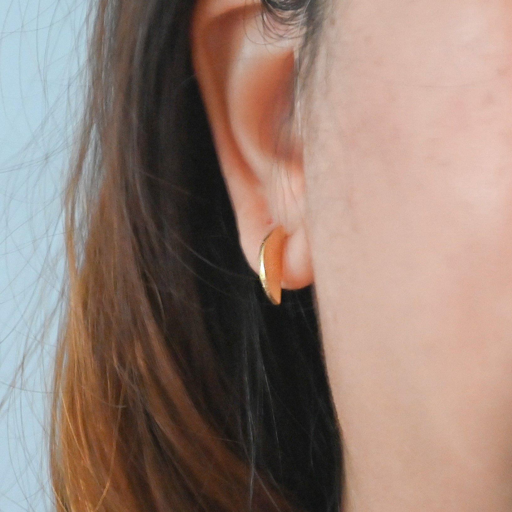 A close-up of a person's ear wearing a small gold crescent-shaped hoop earring against a light blue background.