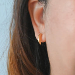 A close-up of a person's ear wearing a small gold crescent-shaped hoop earring against a light blue background.